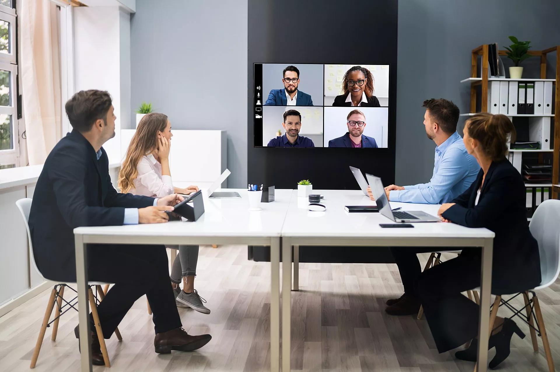 Cuatro colegas se sientan en una mesa de conferencias rectangular y hablan con cuatro colegas por video.