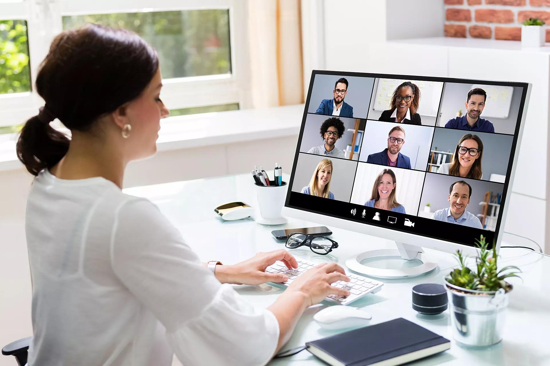 A woman, sitting in an office, types on her keyboard while on videoconference meeting