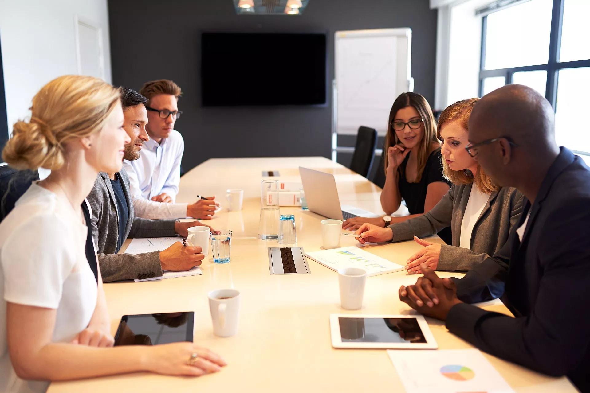 A conference room with a screen at the back and a group of people sitting at the table.