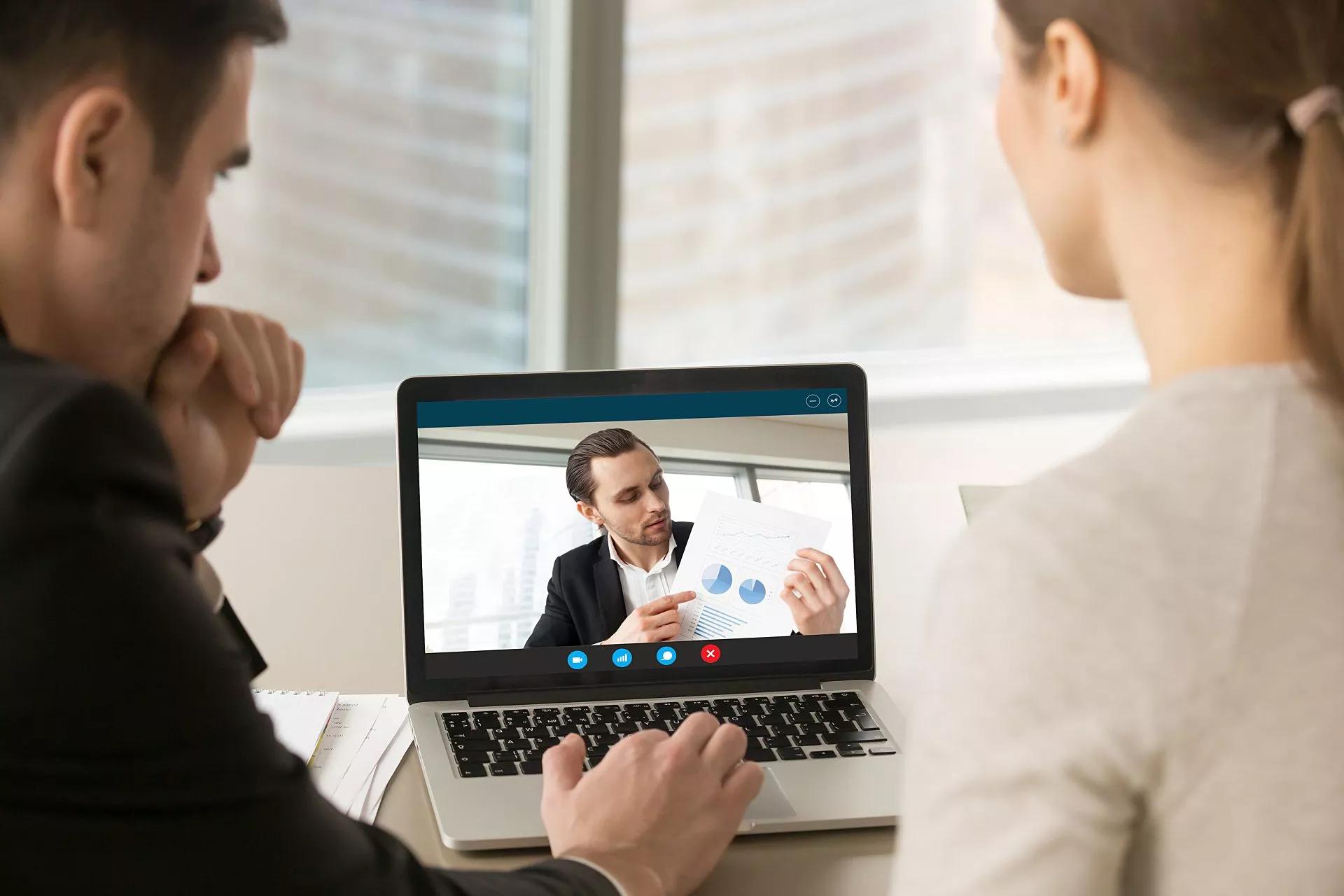 Two colleagues sit in an office and use a laptop to speak to another employee on video.