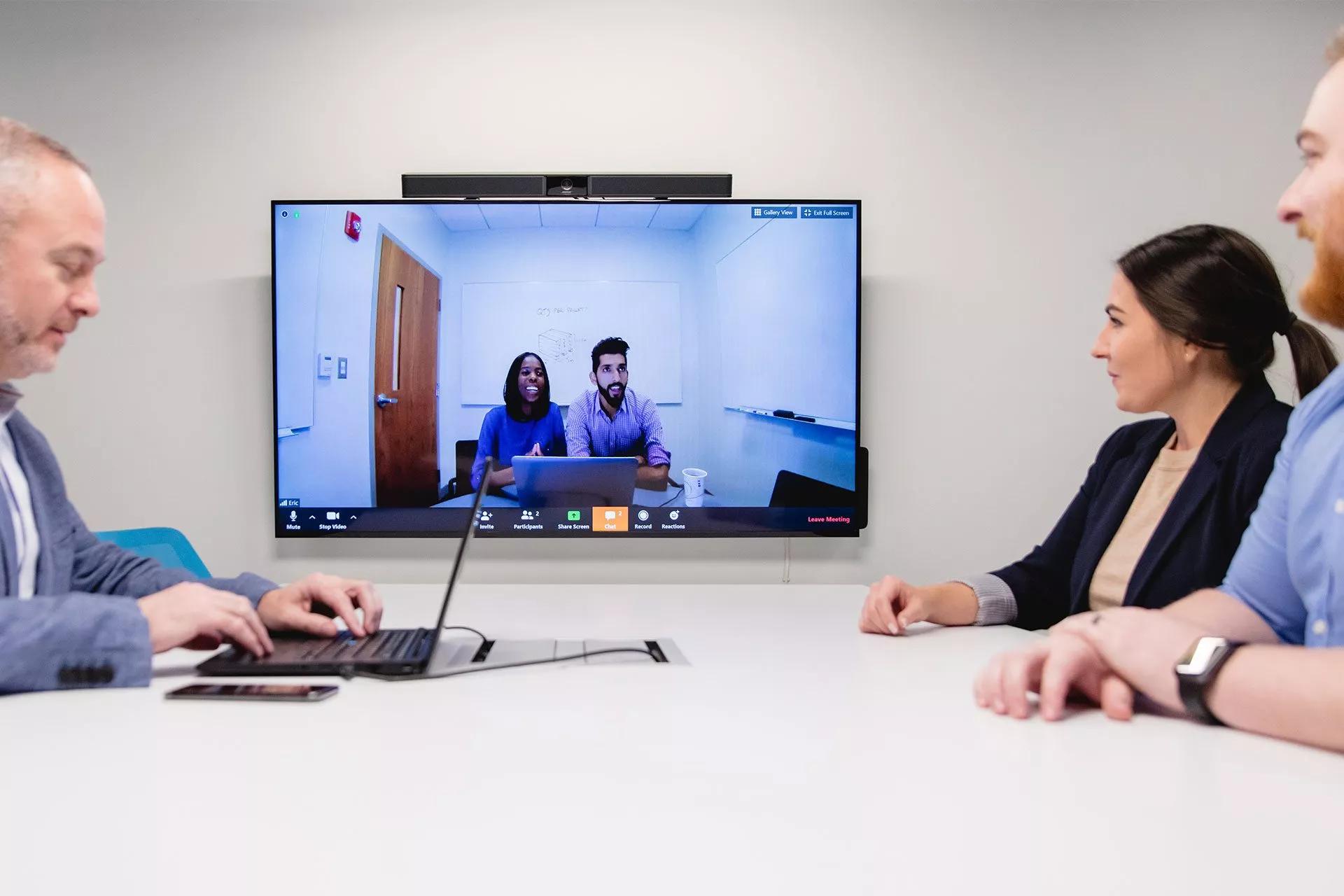 A group of employees sits in a videoconference meeting and view a screen