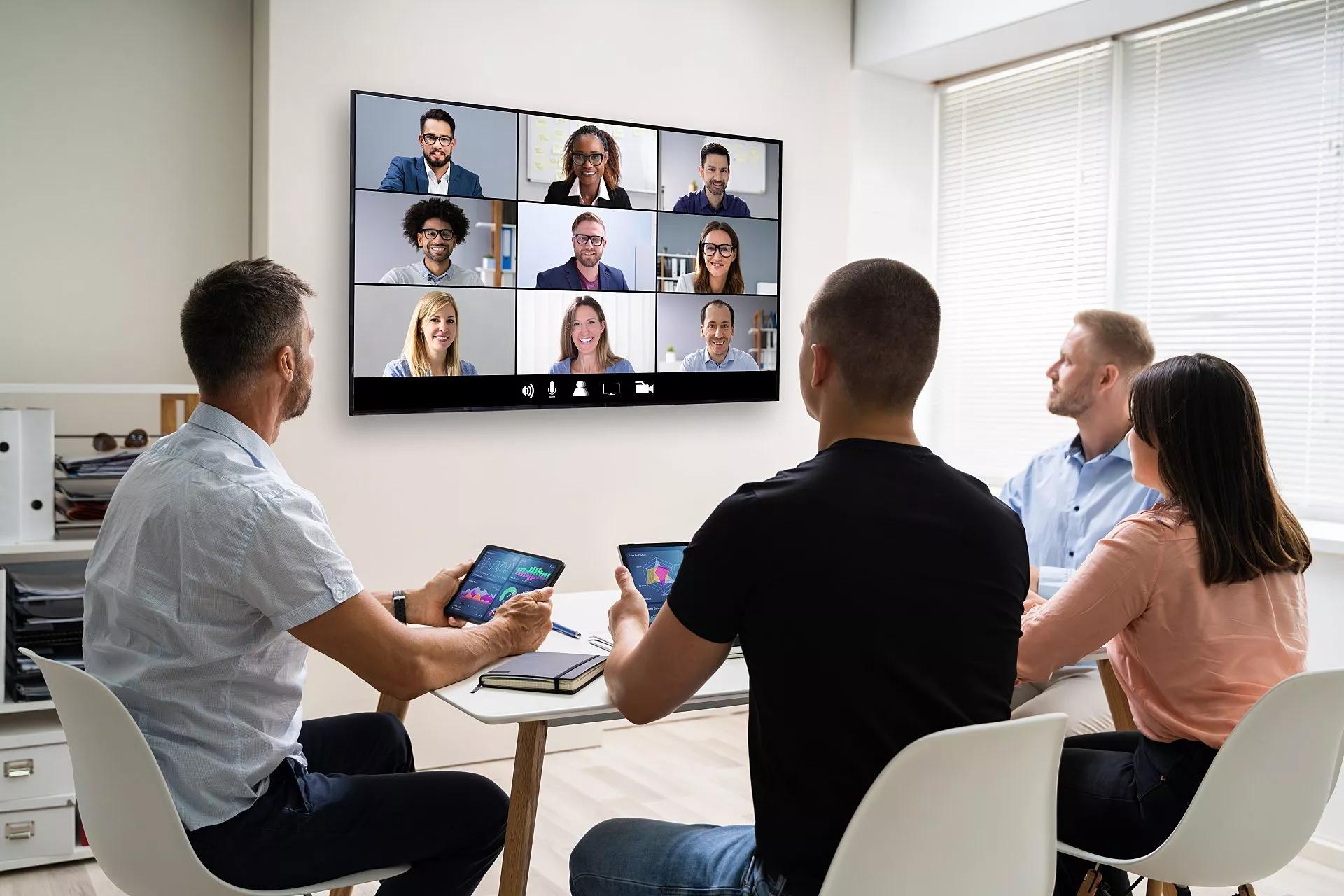 A team sitting around a conference room table interacts with remote colleges via videoconference