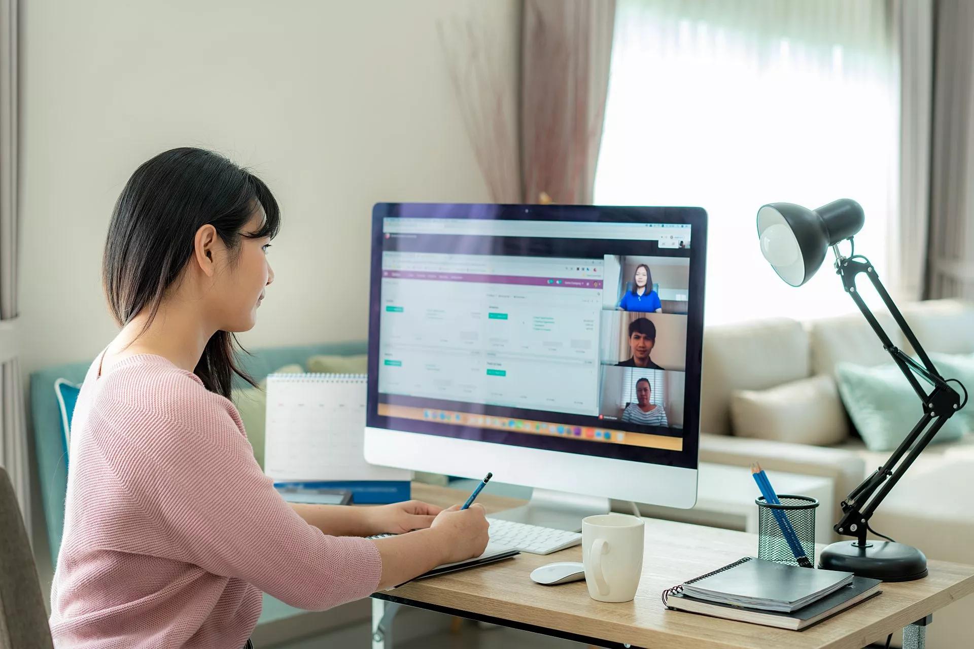 Frau, die von zu Hause aus ein Meeting auf einem großen Monitor am Schreibtisch hat.