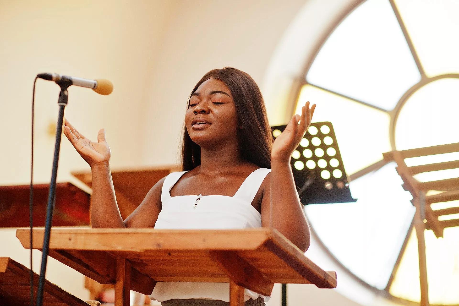 A young woman sings at church with hands held out in prayer.