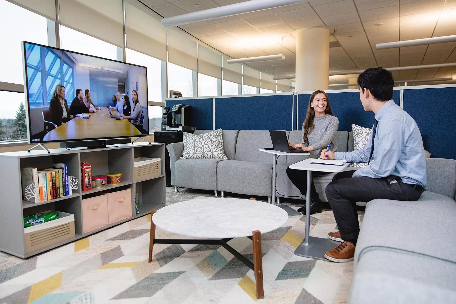 Dos empleados sentados en una oficina de espacio abierto se conectan con un equipo a través de una videoconferencia.