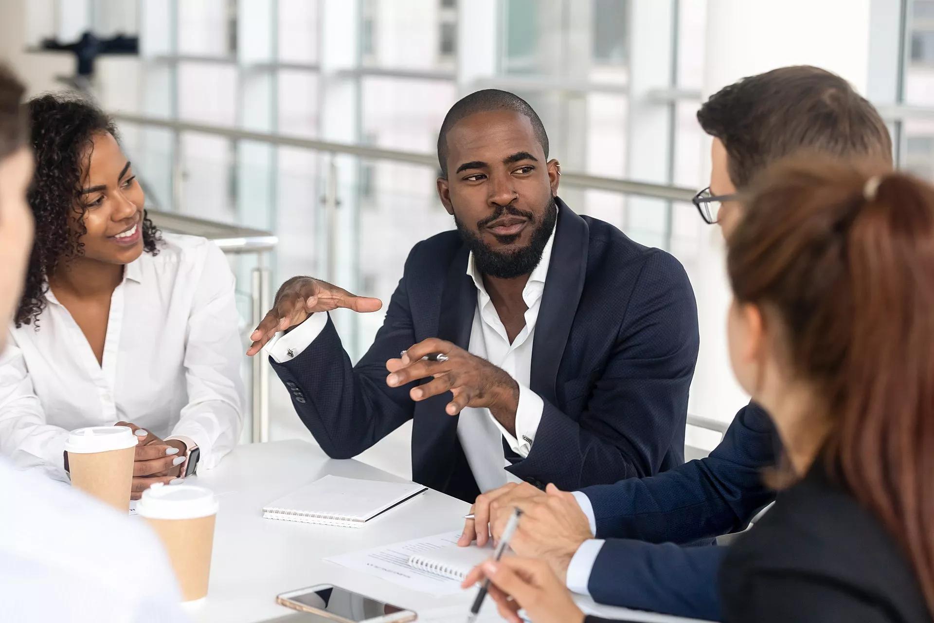 Five workers sitting at a conference table talking to each other.