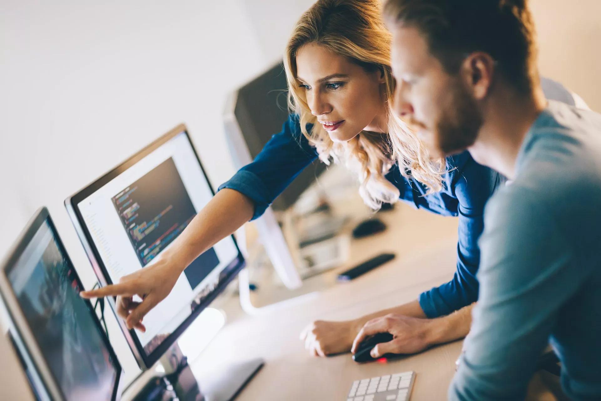 Eine Frau unterhält sich mit einem männlichen Kollegen an seinem Schreibtisch und zeigt auf den Computermonitor