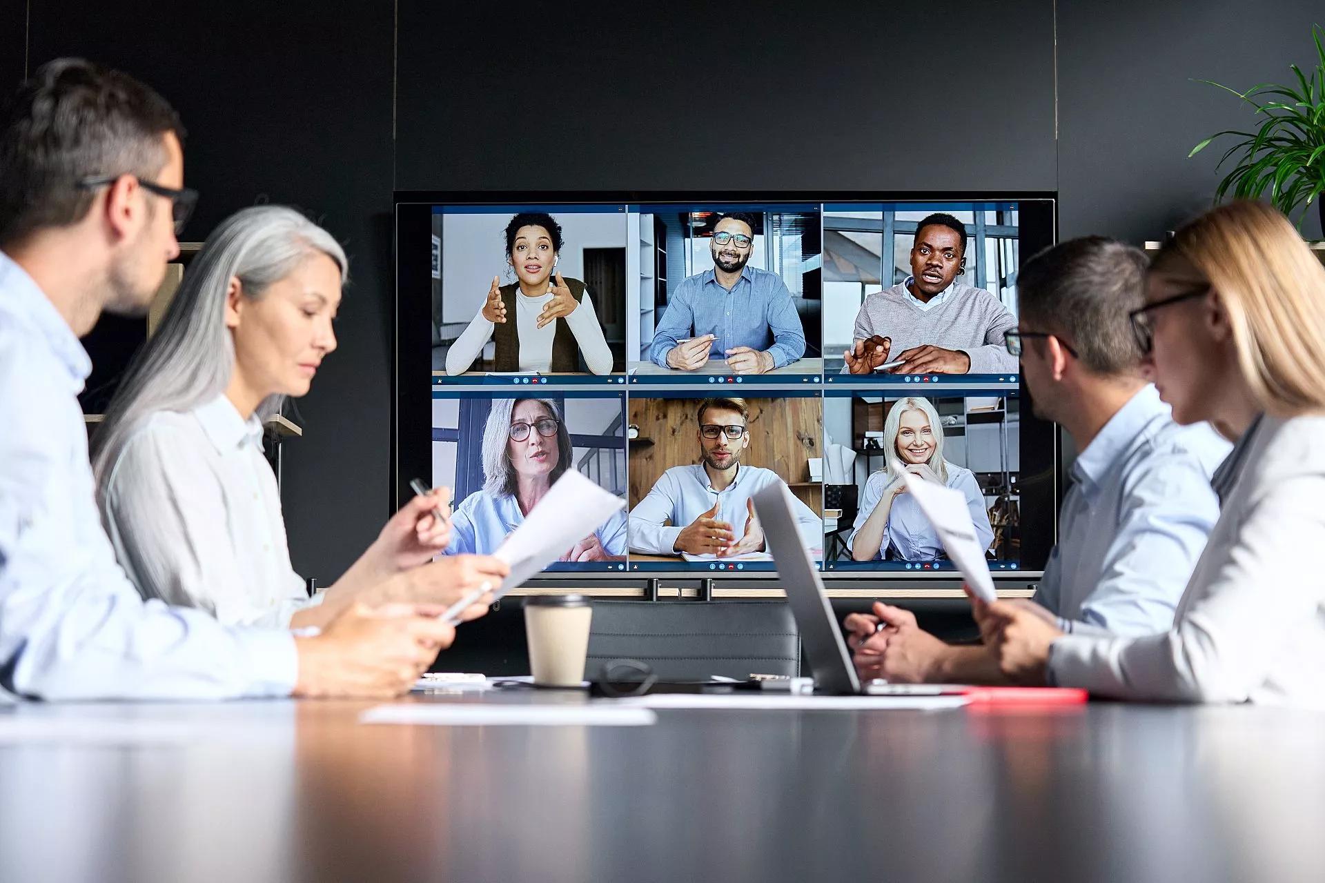 Employees meeting around a table with 6 people on video conference.