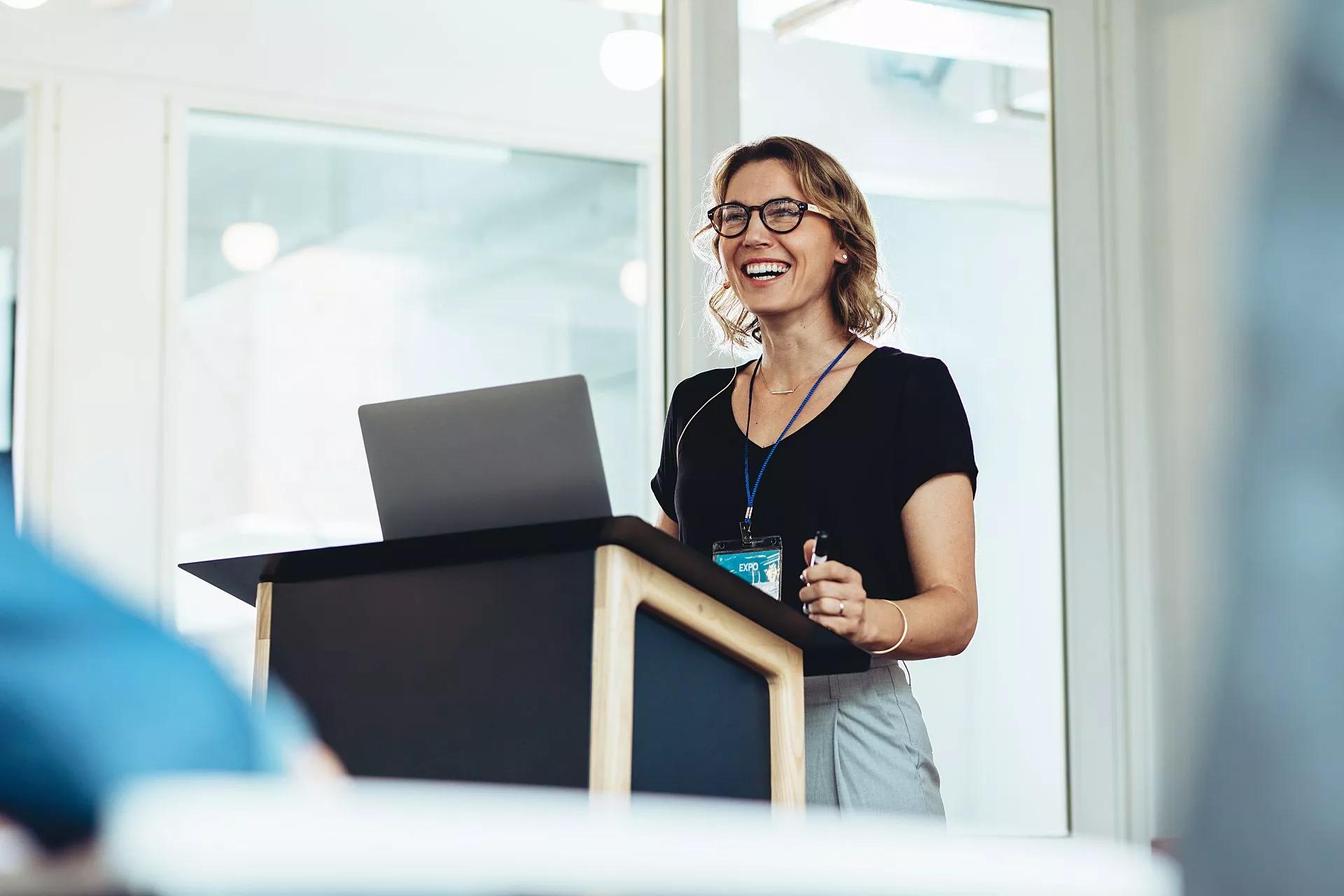 Una mujer de pie frente a un atril con su laptop hace una presentación a los asistentes a una conferencia